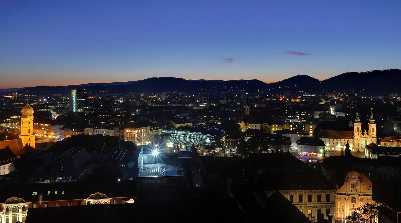 Paar steht auf dem Schlossberg in Graz neben dem Uhrturm und blickt über die roten Dächer der Altstadt