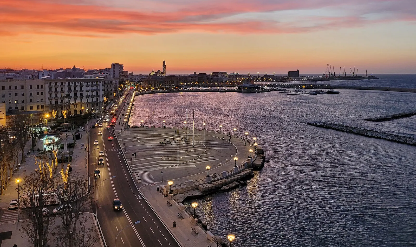 Una coppia passeggia sul lungomare di Bari al tramonto, con il mare a destra e i lampioni accesi
